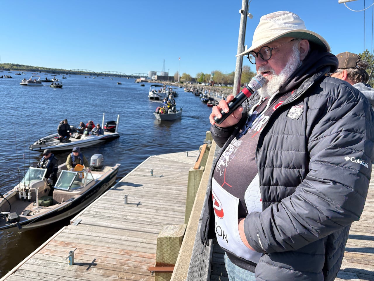 A man in a puffer jacket holds a microphone, overlooking a busy riverfront with many small boats in it.