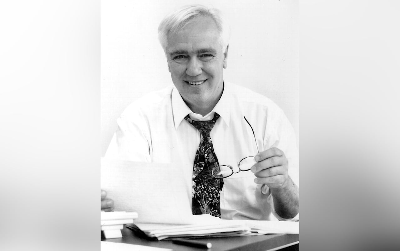 A black and white photo of a man in a white shirt and black tie holding a pair of eyeglasses and looking over papers on a desk while smiling