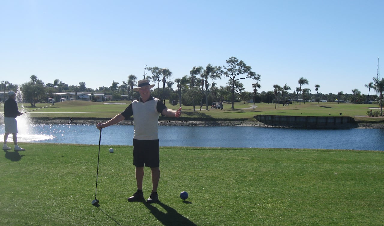 A man wearing a black and white t-shirt, black shorts and a sunhat holds a golf club in one hand and gives a thumbs-up gesture with his other hand while standing on a gold course.