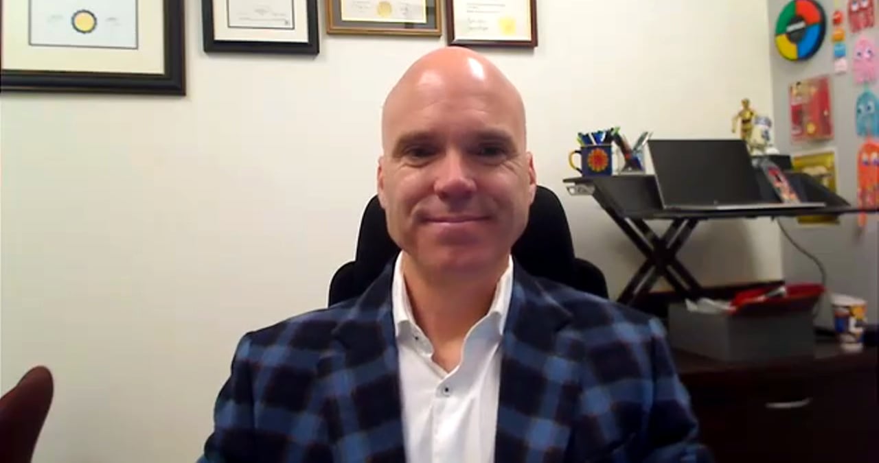 A bald white man sits in his office. He wears a white shirt and a black and blue checkered jacket.
