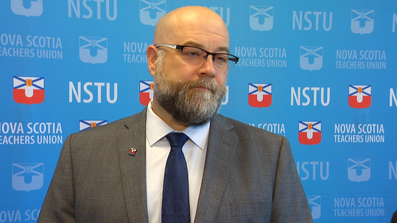 A bald white man wears glasses and a grey suit with a navy blue tie. He stands in front of a bright blue backdrop that says 'NSTU' and 'Nova Scotia Teachers Union.'