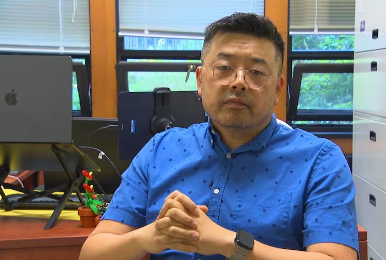 An Asian man sits in front of his desk with his hands folded. He wears a blue collared shirt and glasses. He has a short haircut.