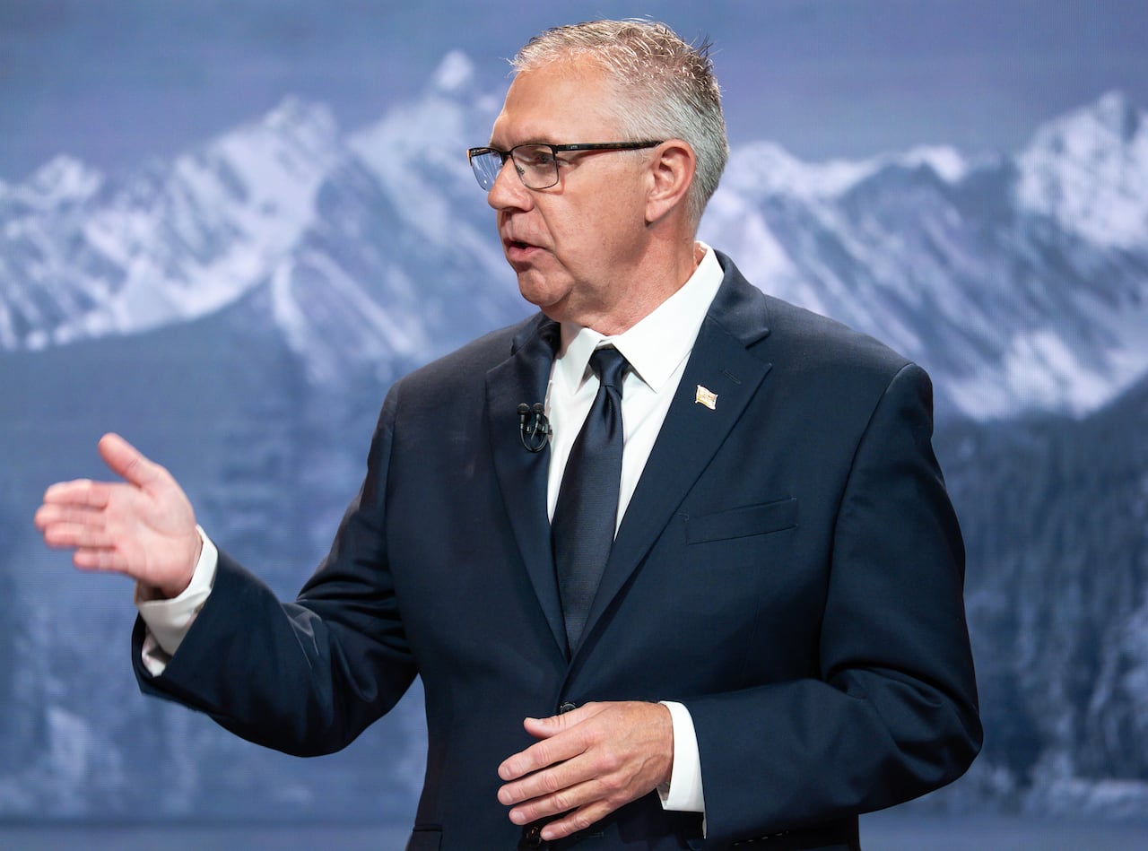 A white man wearing a suit is seen in front of a mountain background.