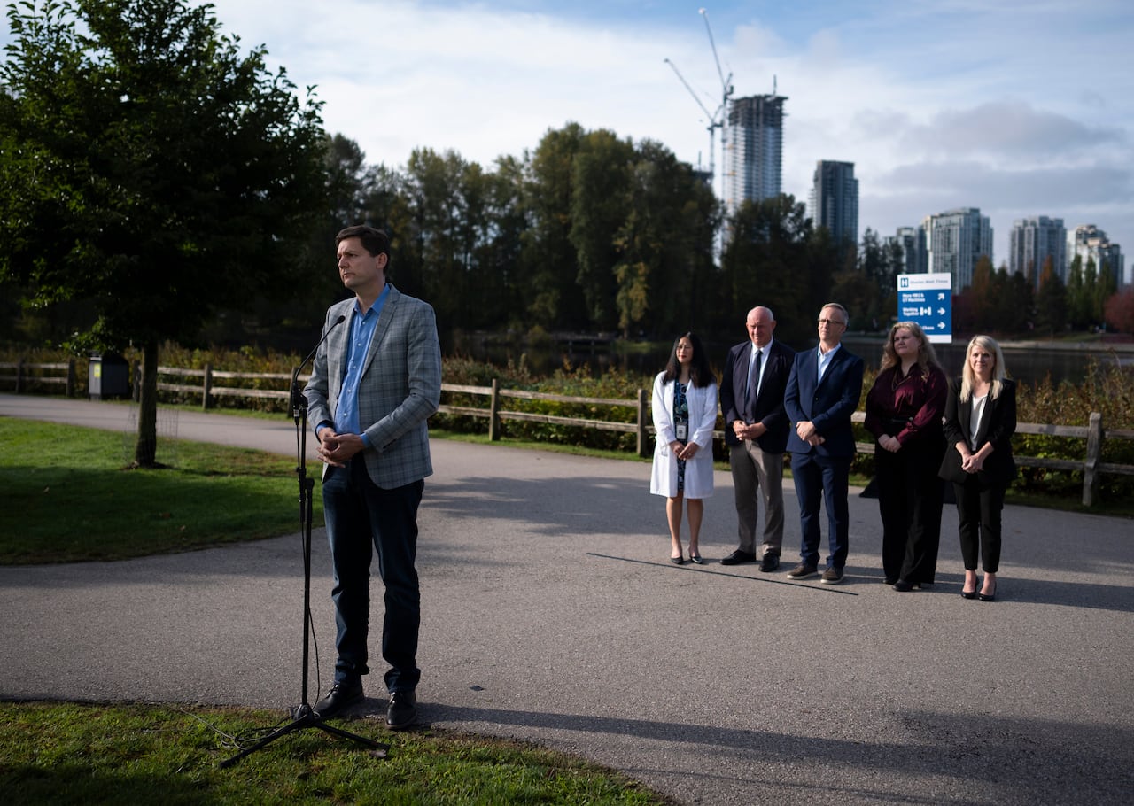 A tall white man is seen at a mic outdoors, with a gaggle of people behind him.