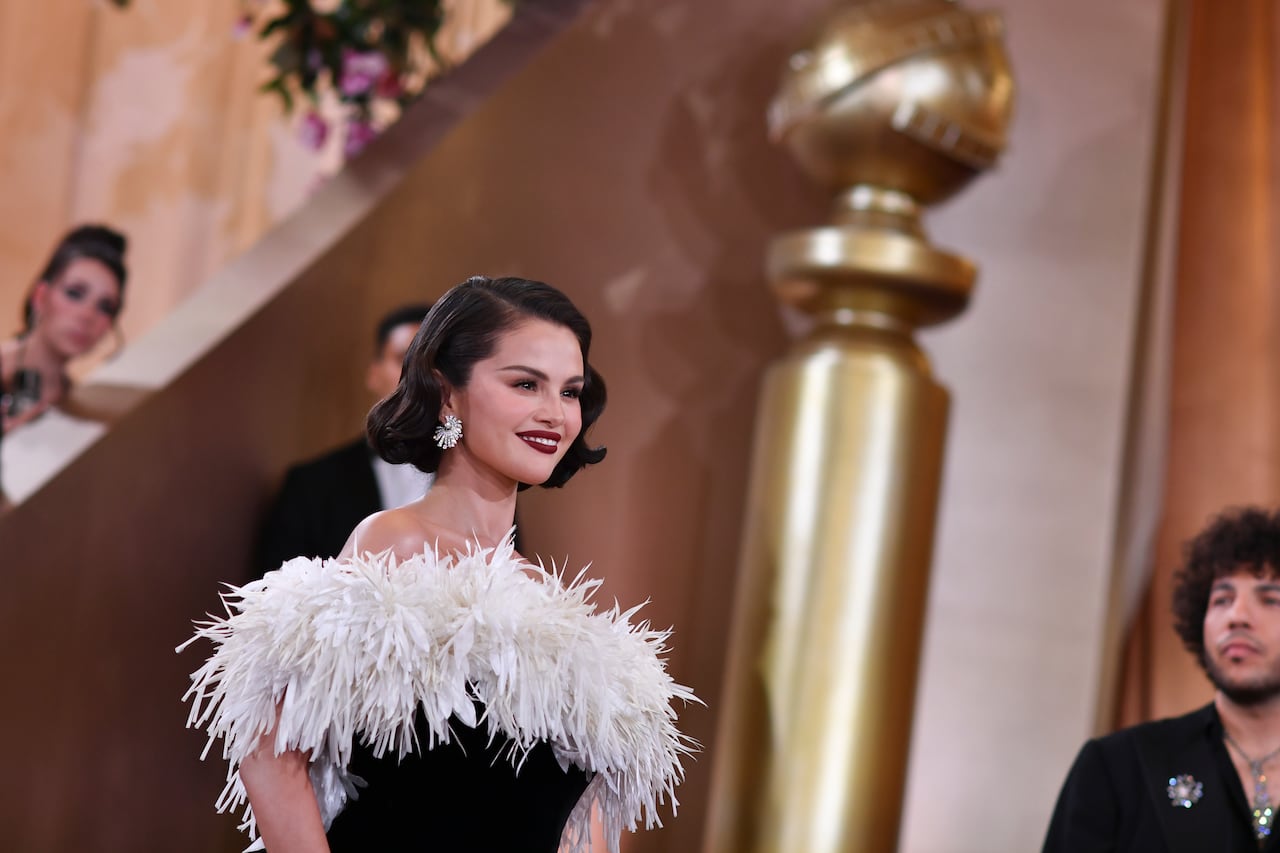 A woman in a black dress with white feathers smiles as she walks down a red carpet with gold embellishments.
