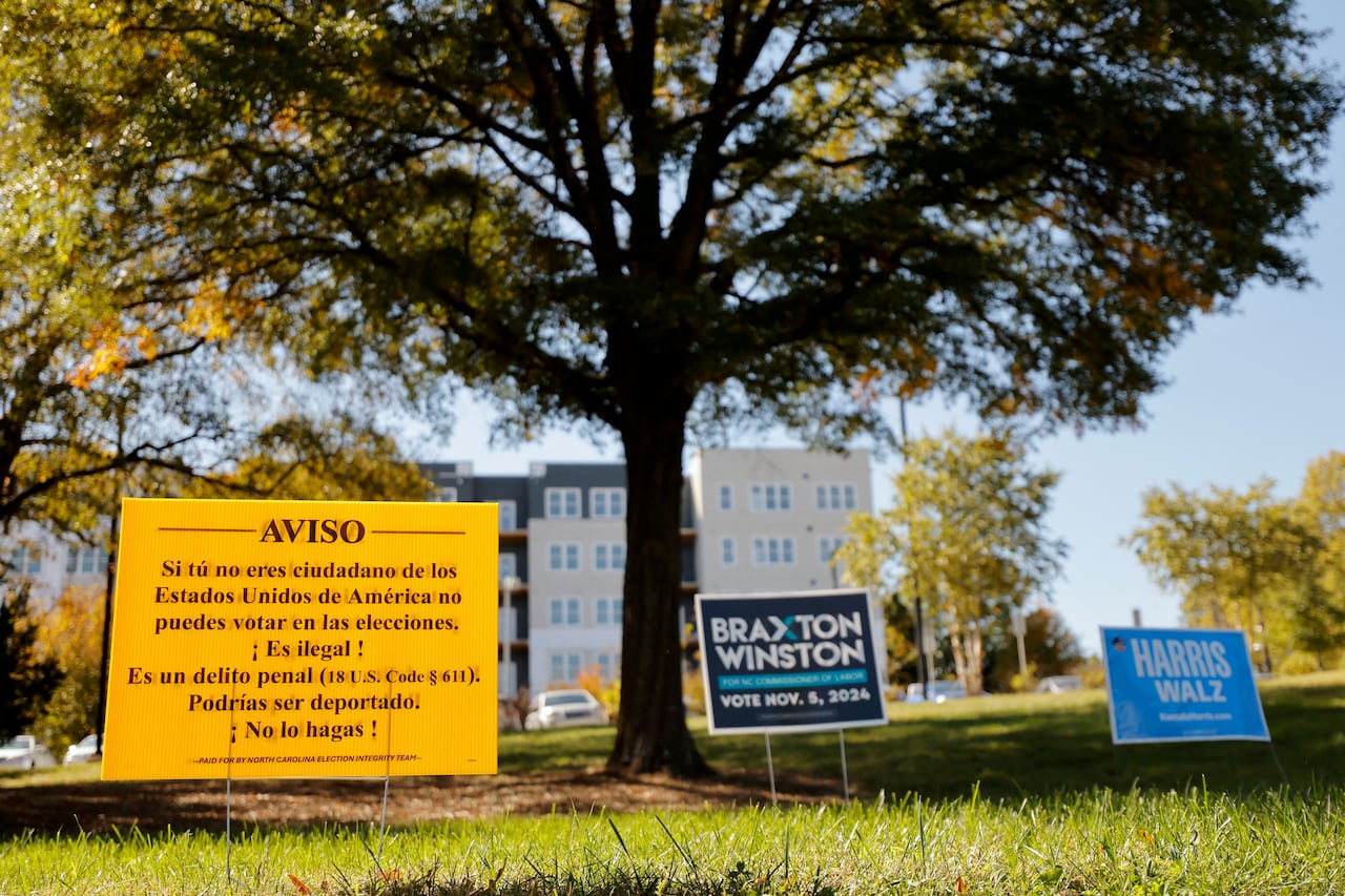 A yellow sign with text stands in some grass in front of buildings.