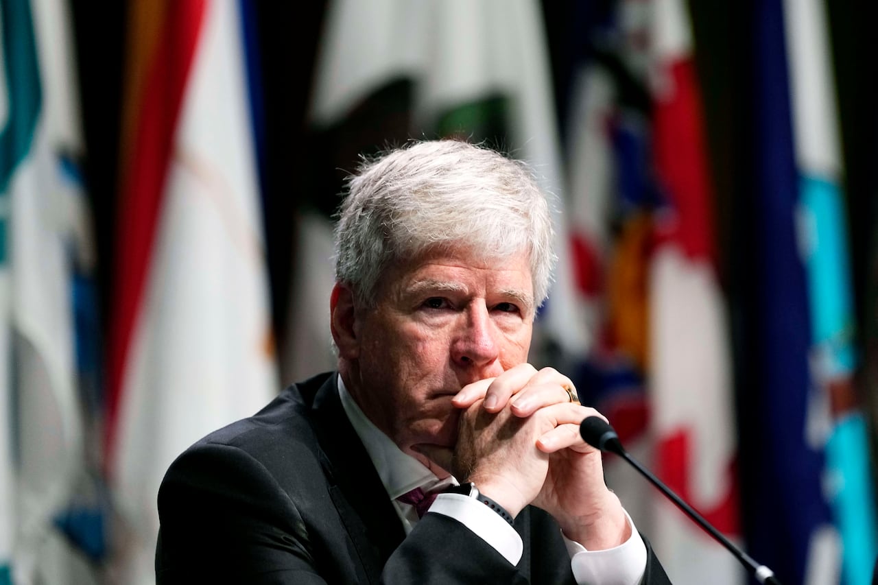 A man sits at a microphone with his hands clasped. There are flags in the background.