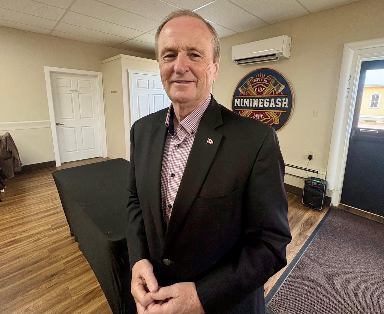 A man in a dark suit stands in a yellow room with a sign that reads 'MIMINEGASH' behind him.