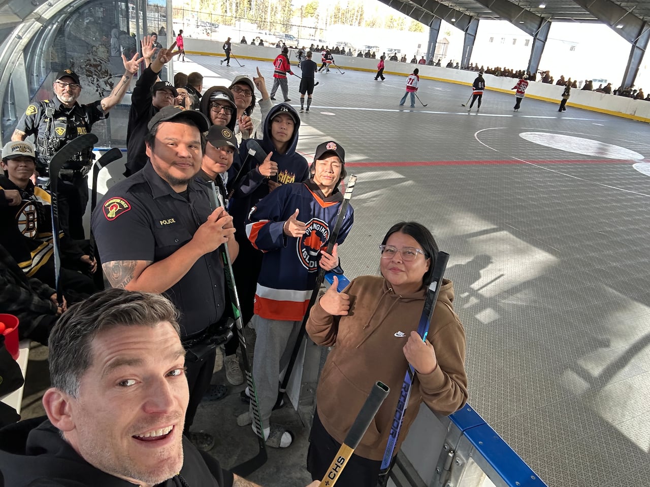 A man takes a selfie with youth holding hockey sticks with a large rink in the background.