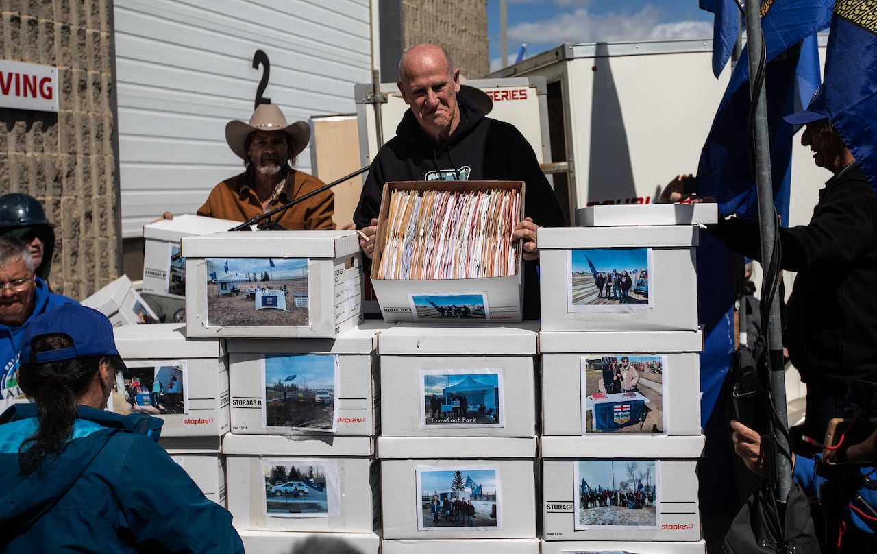 A man in a black hoodie stands smiling behind a stack of boxes, with people and Alberta flags seen beside him.