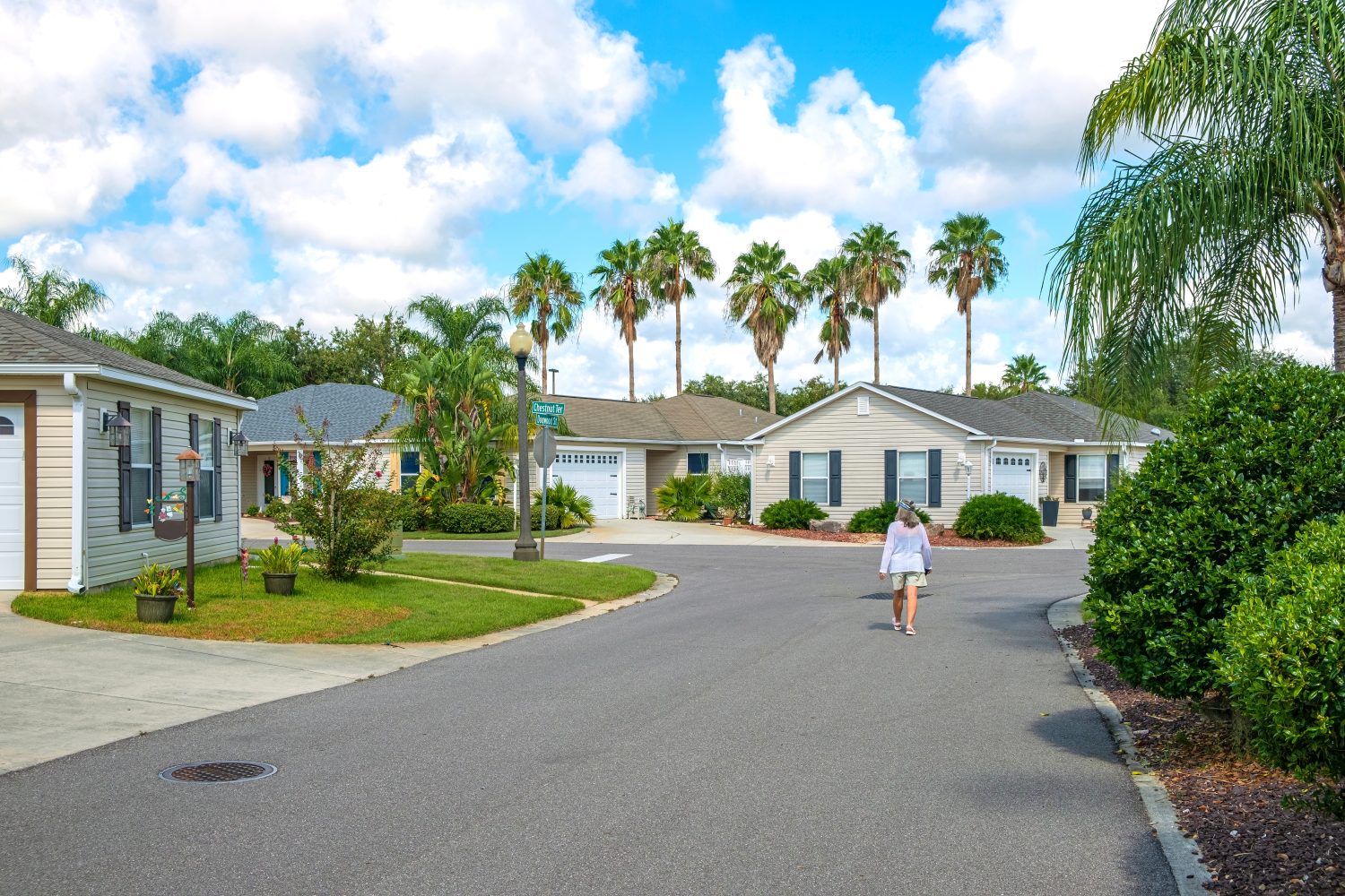 An overhead view of a well-maintained residential street in a warm climate, with single-story beige houses featuring dark shutters and attached garages. Lush green lawns and numerous palm trees line the street, which curves gently to the right. A person in a white top and shorts walks away from the viewer down the asphalt street. The sky is bright blue with scattered white clouds.