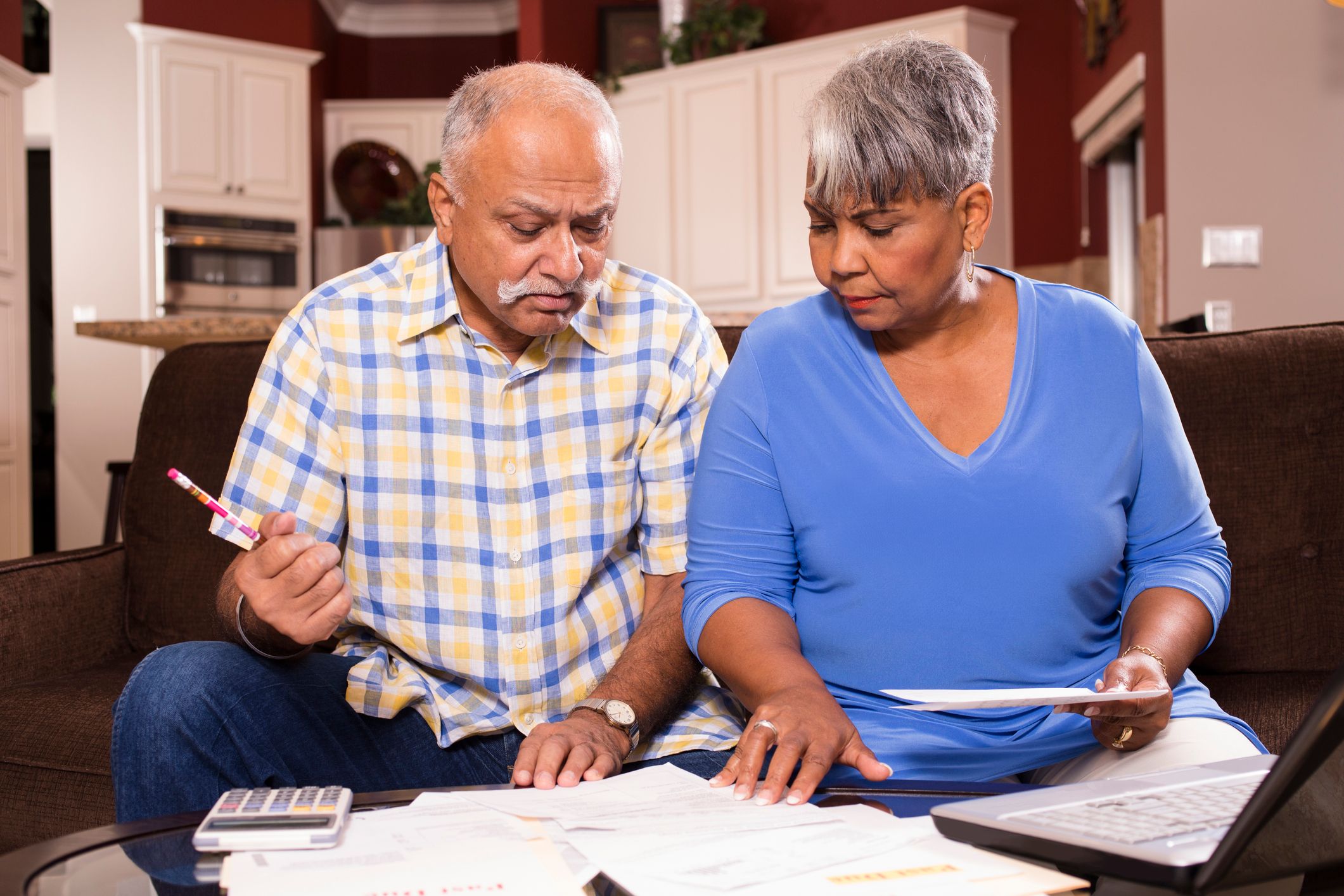 A couple is sitting on a couch, reviewing bills and financial statements set on a table in front of them.