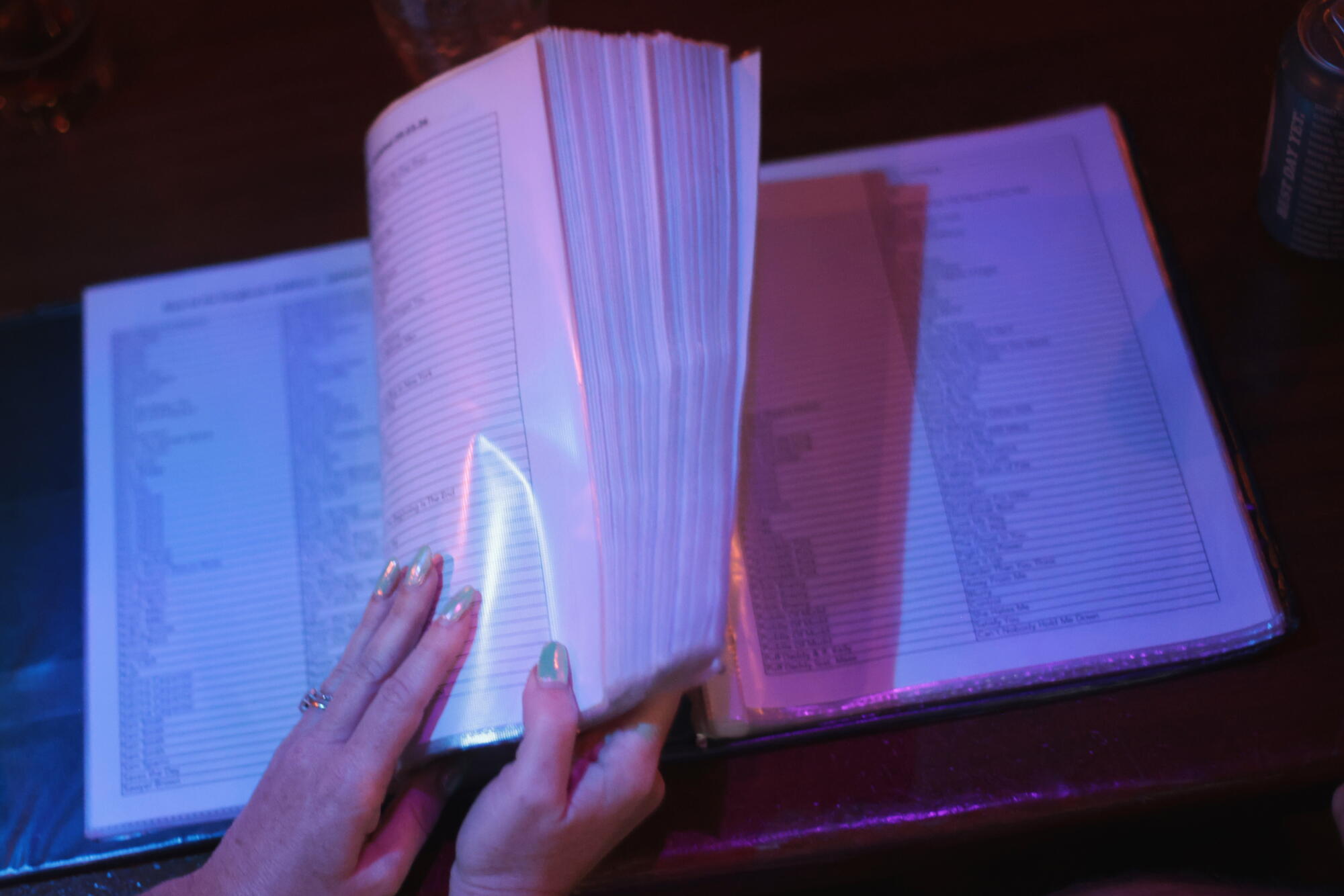 Hands with silver nail polish flip through a large karaoke book with laminated pages.