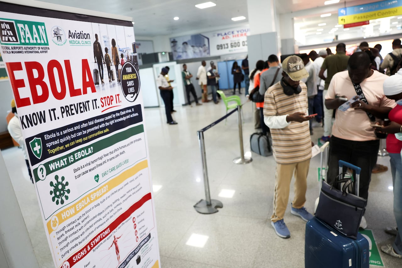 People in line at an airport, looking at their phones as they stand next to their suitcases. A large white, vertical banner bears the word "Ebola" in big red letters along with other text explaining the signs, symptoms and how to prevent transmission