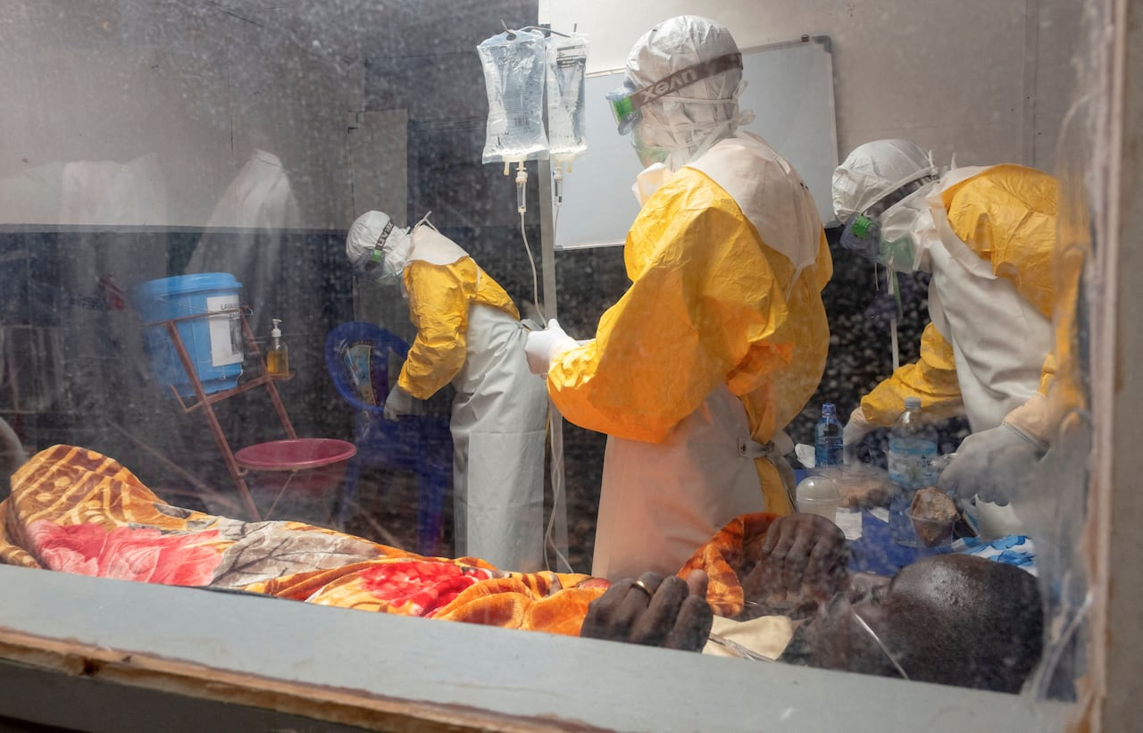 Medical workers clad in yellow protective suits and white hoods, with goggles over their faces, are seen working in a treatment room, behind clear plastic, while a sick man lays on a bed, breathing through an oxygen tube.