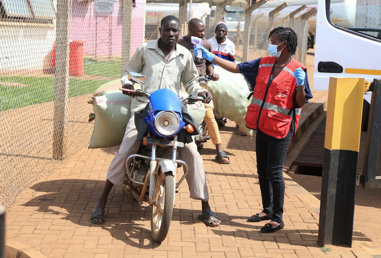 A woman, wearing a bright red vest, stands beside a man on a motorbike and holds a temperature scanner to his size of his head.