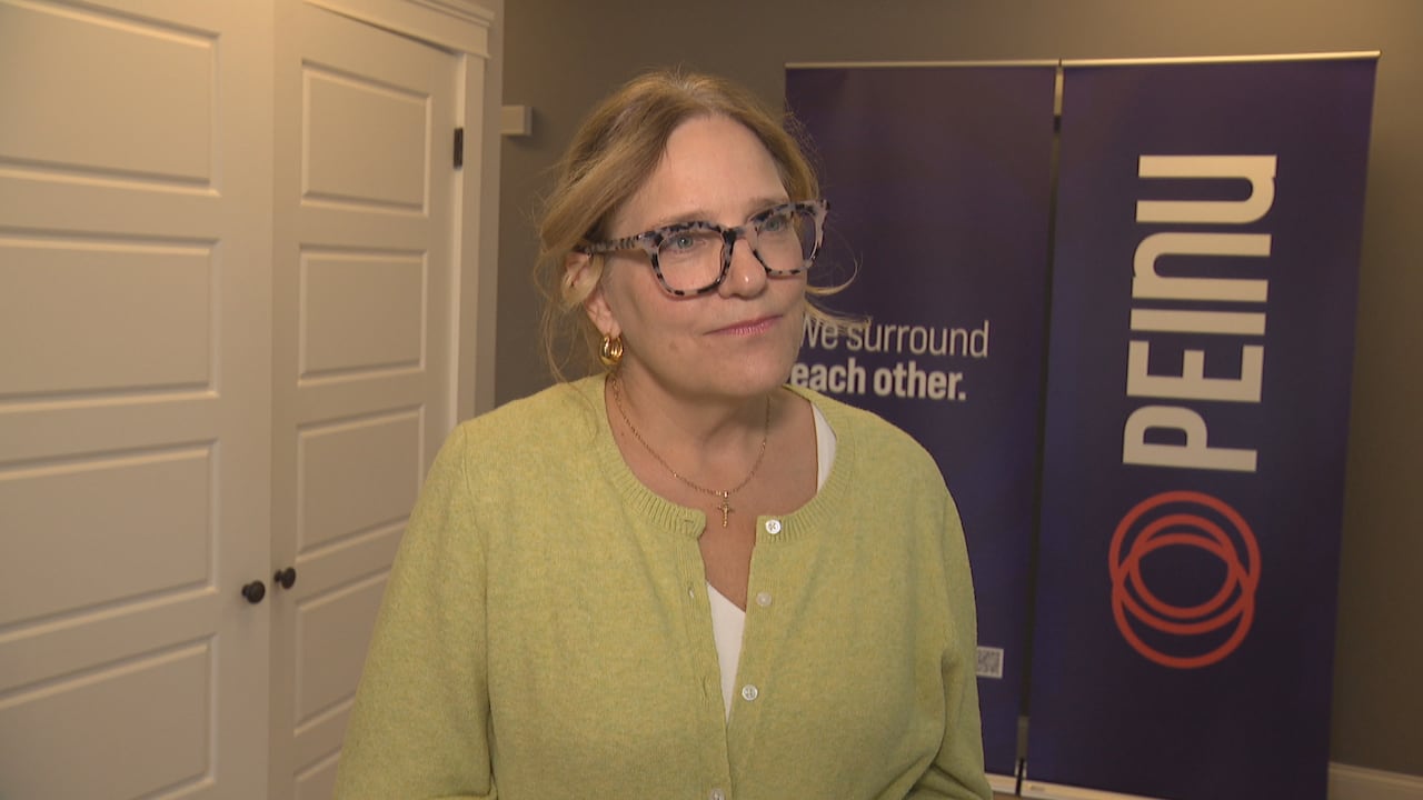 A woman in glasses and a light green top stands near two white doors and in front of a sign that says 'PEInu' in white letters.