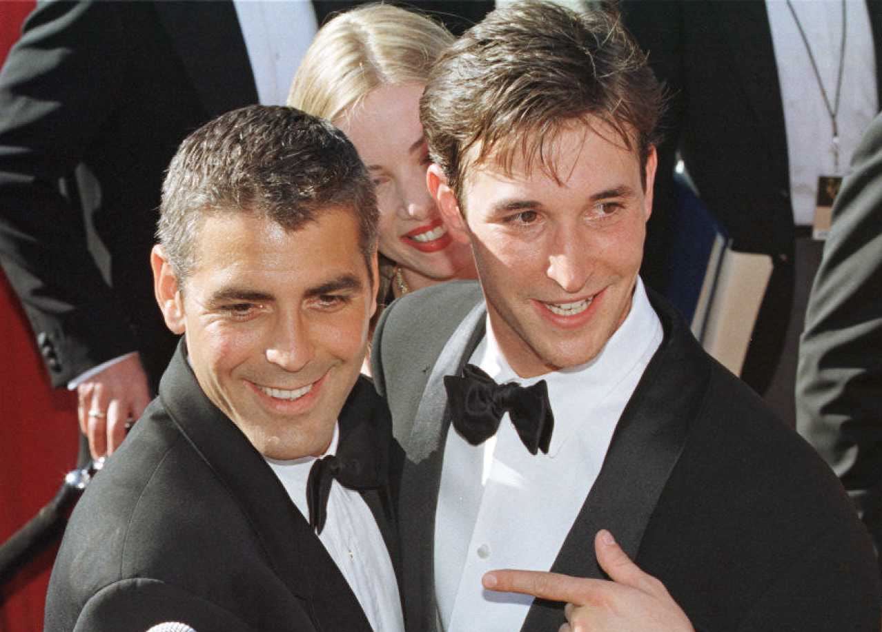 Two men in suits pose together, smiling, with the one on the right pointing to the other one.