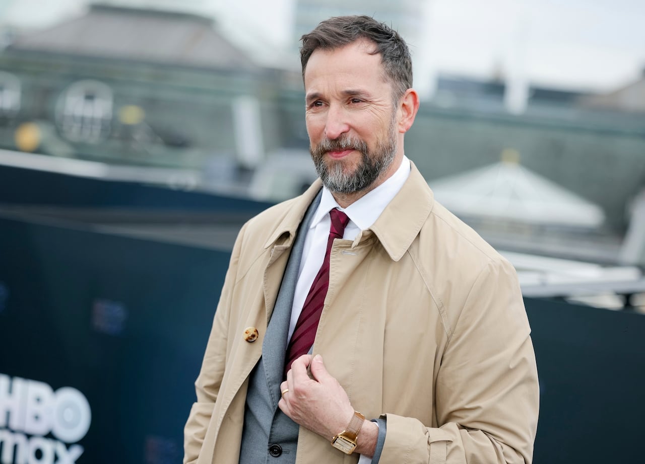 A man in a beige coat over a grey suit poses for a photo.
