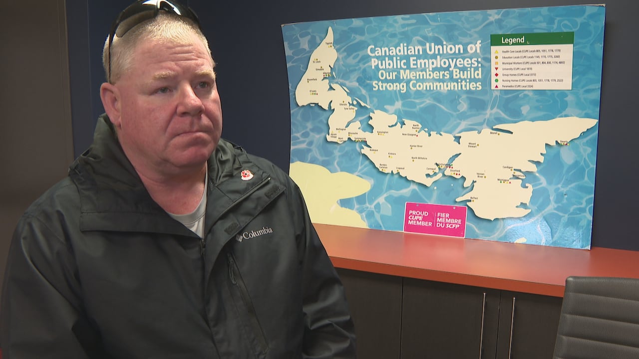 A man in a dark jacket stands near an image of Prince Edward Island with a blue background.