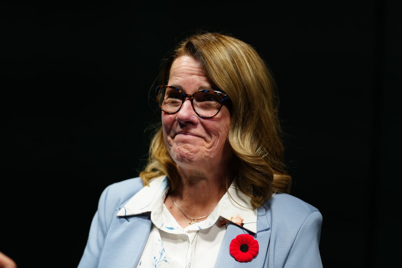 A woman wearing glasses and a light blue blazer smiles.