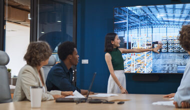 Four people in a conference room watch a presenter point to a warehouse scene on a large display.