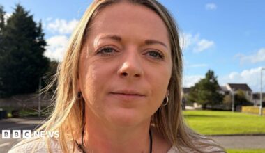 Close-up portrait of a woman with a nose ring and hoop earrings standing outdoors on a sunny day, with a grassy area, trees and houses blurred in the background.