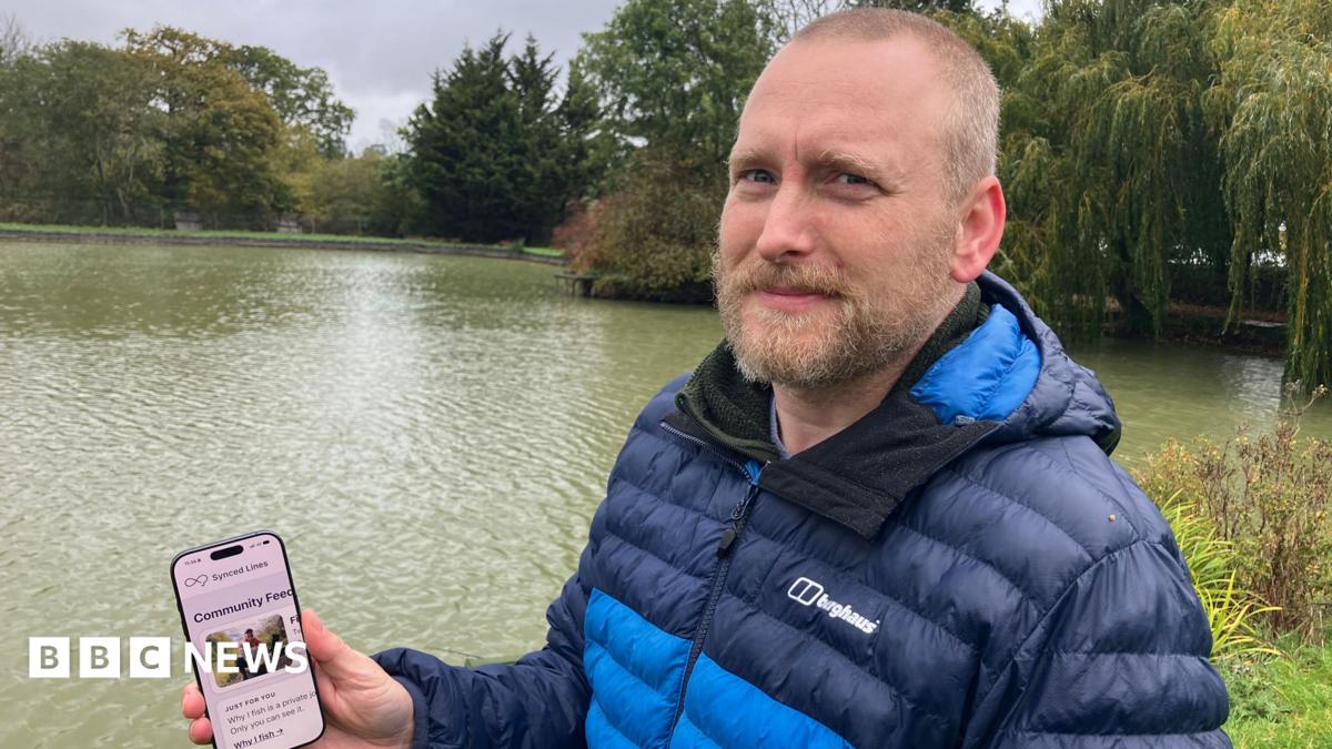 A man in a navy and light blue puffer jacket holds up a phone. He is standing in front of a body of water.