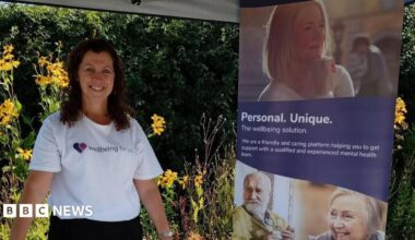A woman with dark hair and wearing a white t-shirt with a wellbeing logo stands next to a display stand featuring several faces and a caption which reads: "Personal, Unique." There is greenery in the background.