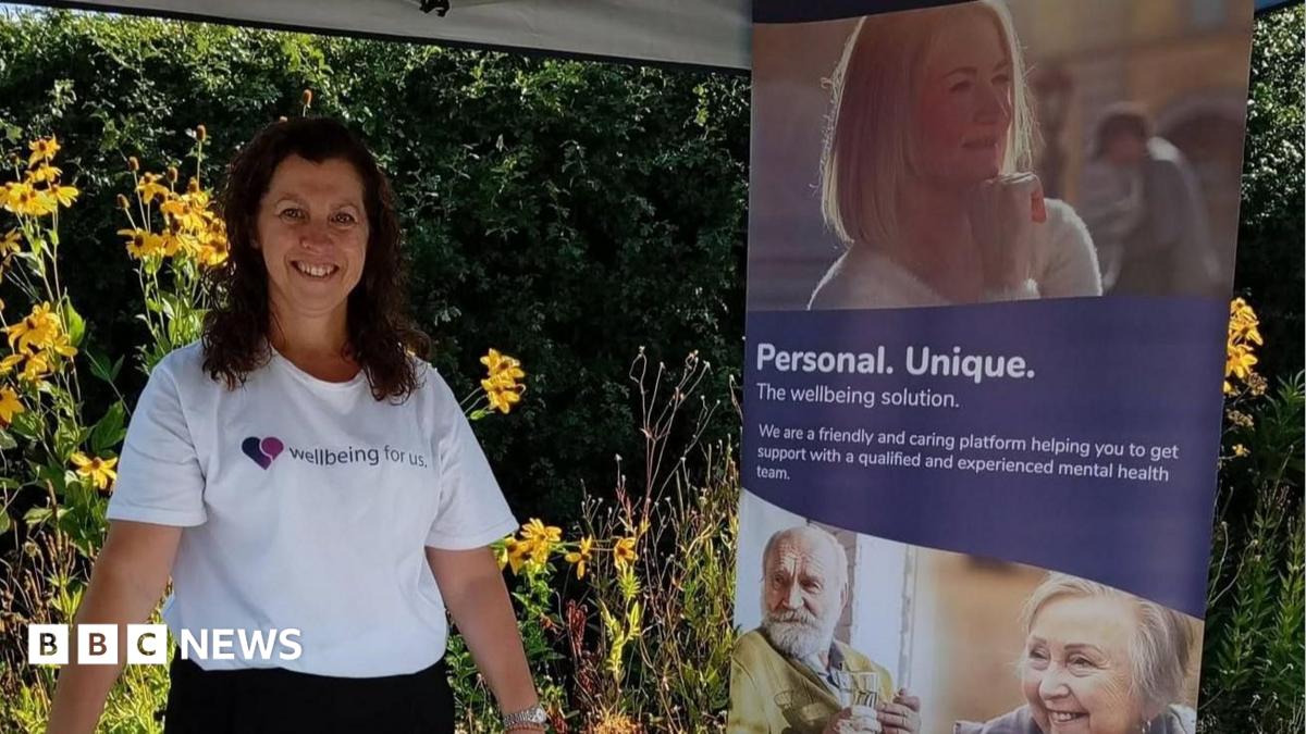 A woman with dark hair and wearing a white t-shirt with a wellbeing logo stands next to a display stand featuring several faces and a caption which reads: "Personal, Unique." There is greenery in the background.
