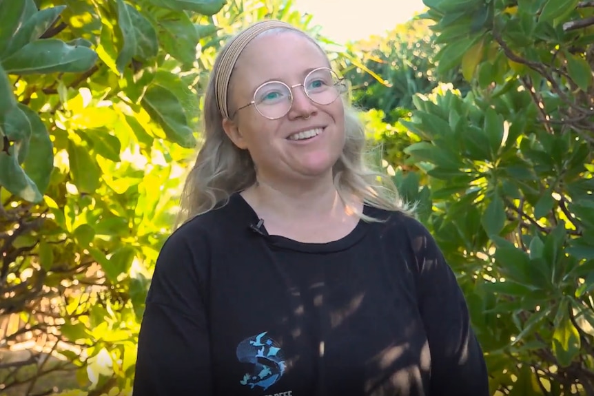 Zerra is a smiling woman wearing spectacles, standing beween two hedges.