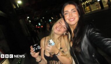 There are two girls in a dark bar smiling at the camera . On the left a blonde girl is wearing a strappy top and holding a drink. She leans into her friend on the left who is holding the camera to take a selfie of the two of them. The girl on the right has long brown hair and is wearing a black leather jacket.