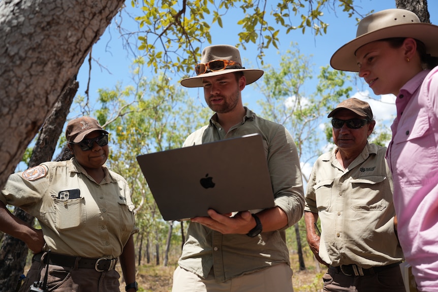 Two uniformed rangers and a pair of researchers in work shirts huddle around a laptop screen as they stand in a national park.