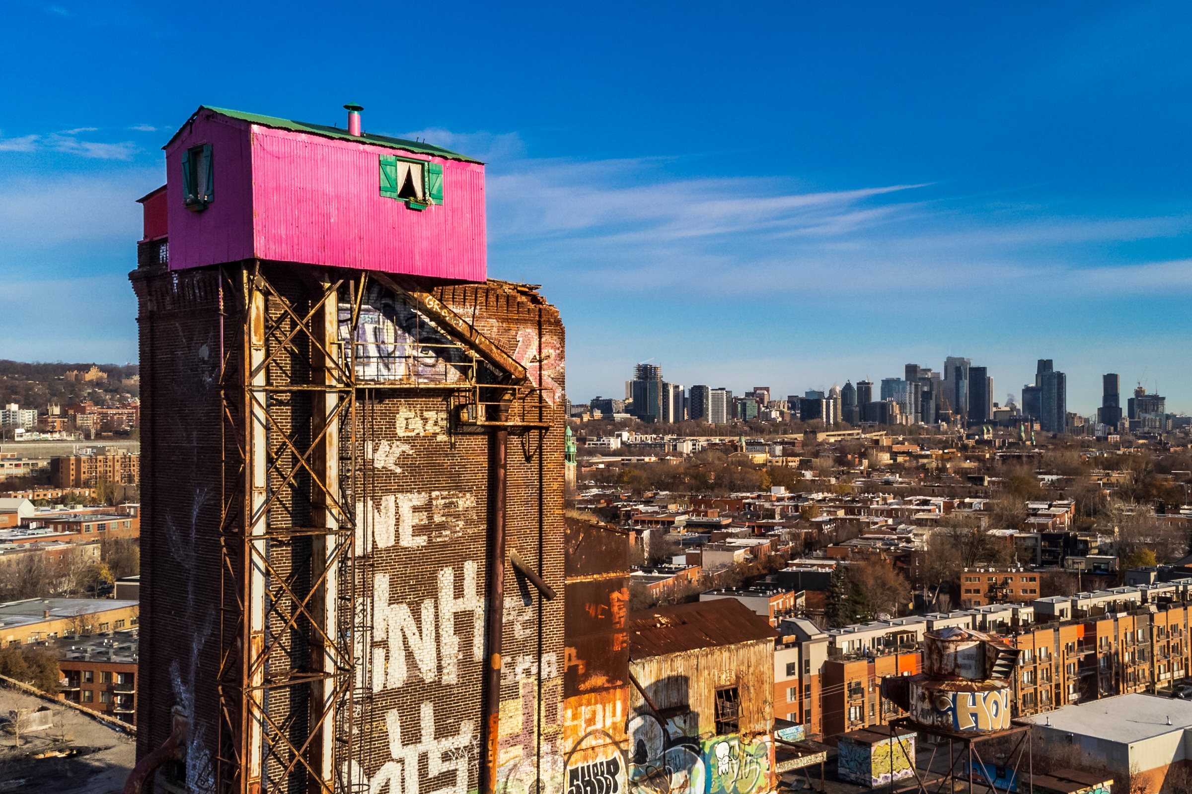 A small pink house atop an industrial tower, with a city skyline in the background.