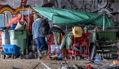 A man approaches a structure in an encampment under an overpass.