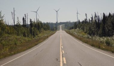 A two-lane highway recedes into the distance, where wind turbines stand on the horizon.