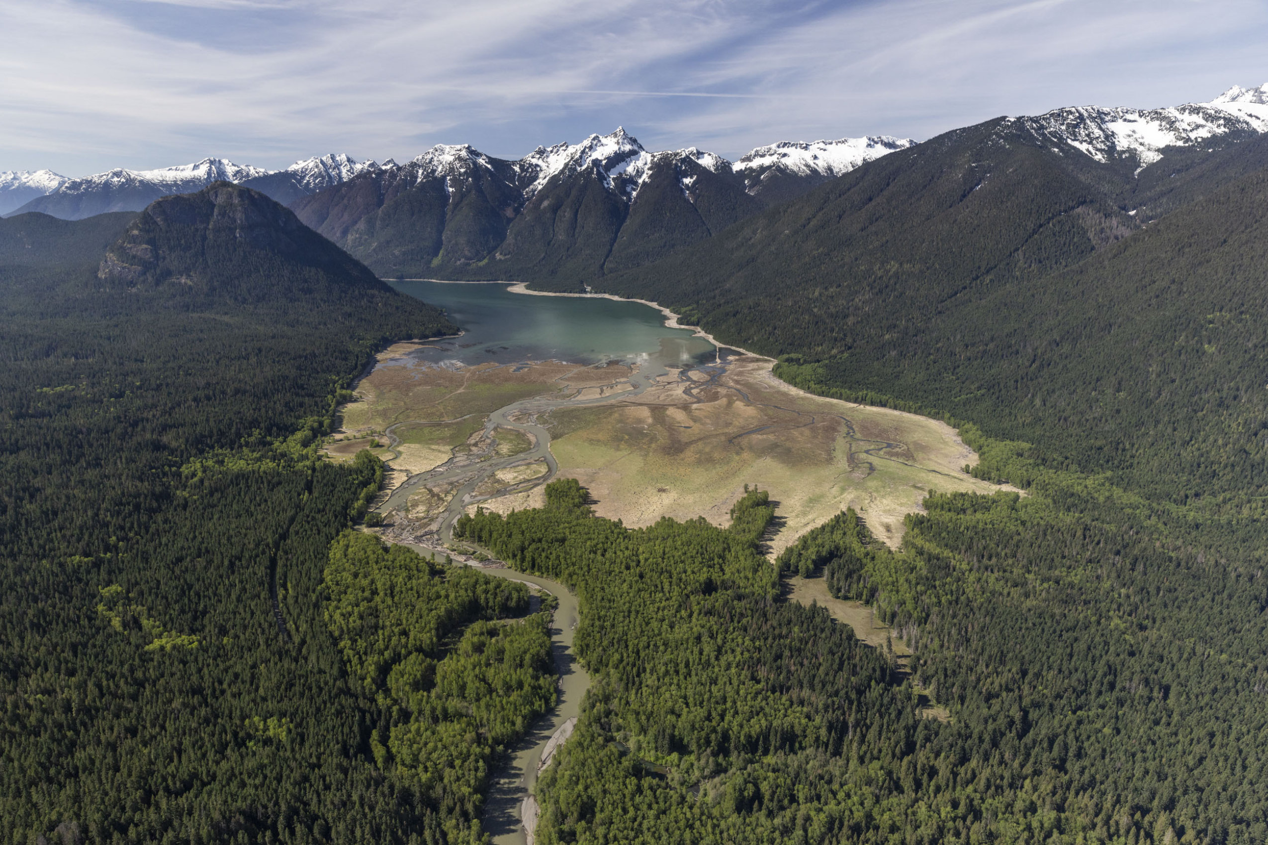 An aerial view of a wilderness landscape, with forested areas, a river and snow-capped mountains.
