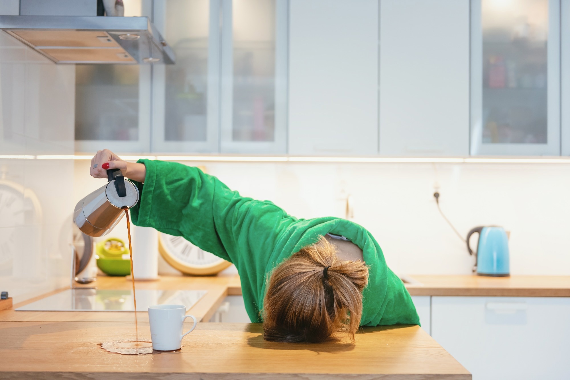 Tired woman sleeping on the table in the kitchen at breakfast. Trying to pour morning coffee into mug but spilling on to the counter