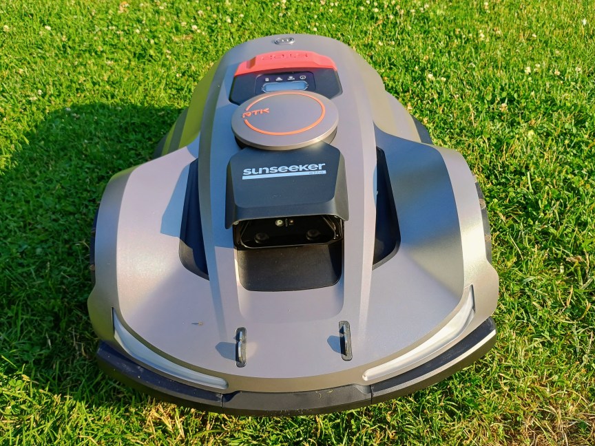 A bronze-colored robot mower by Sunseeker with black and red accents, sits on a bright green lawn.