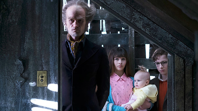 Neil Patrick Harris  as Count Olaf standing in front of Malina Weissman as Violet holding baby Presley Smith as Sunny next to Louis Hynes as Klaus