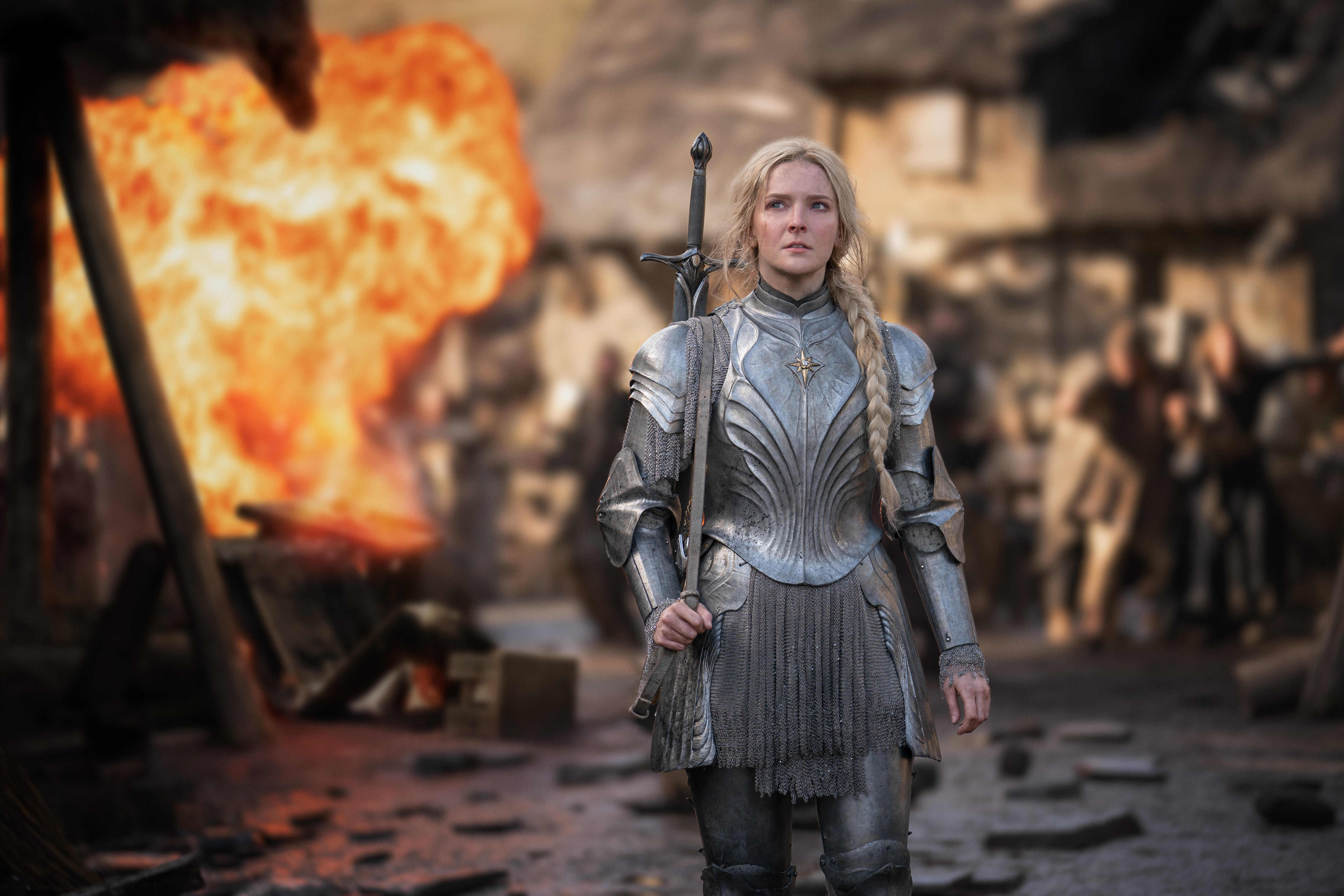 A young Galadriel standing in silver armor with a fiery explosion behind her in a village