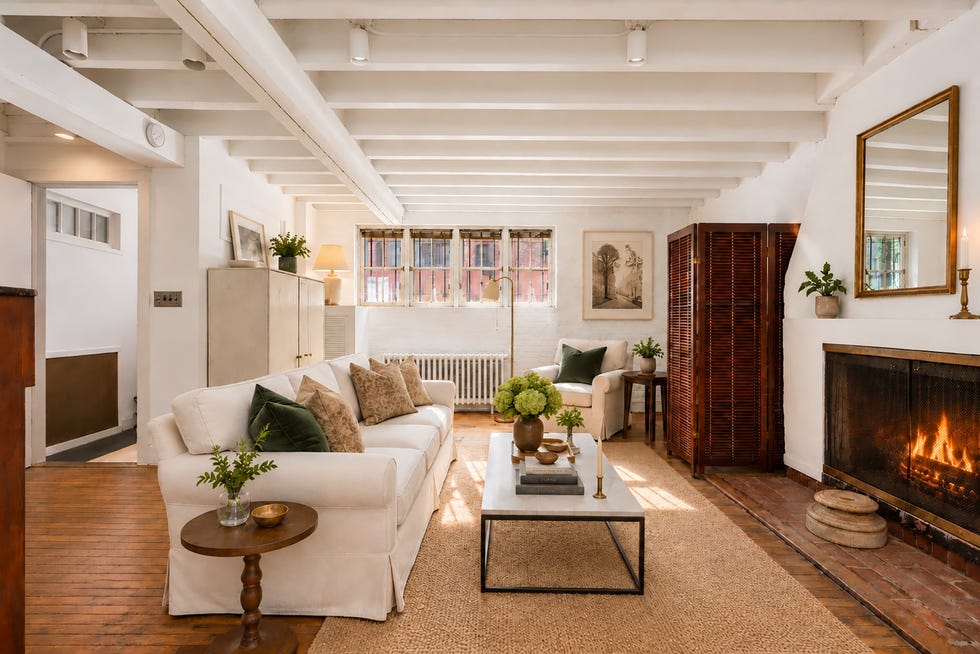Cozy living room with a white sofa, coffee table, plants, art, and a fireplace, featuring warm wooden accents.