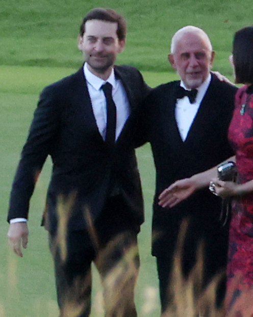 Tobey Maguire in a black suit with a black tie standing next to a man in a tuxedo with a bow tie.