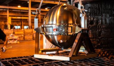 Polished stainless steel spherical probe suspended by lifting straps above a metal furnace tray in an industrial workshop