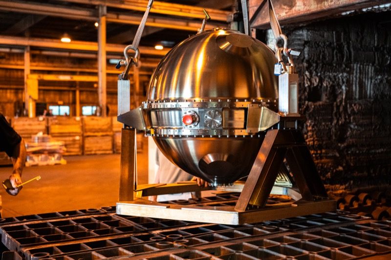 Polished stainless steel spherical probe suspended by lifting straps above a metal furnace tray in an industrial workshop