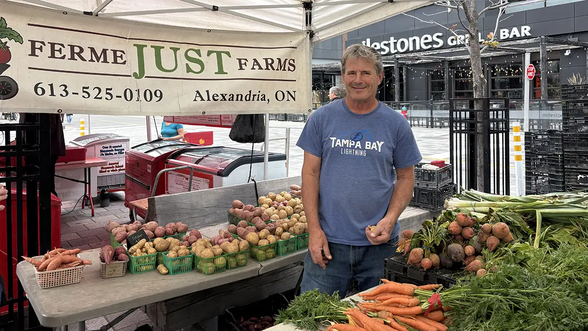 Marc Just standing within his farmers market stand surrounded by his fresh produce. This image provides a personality and context to who is providing input into the relevance of time change among Ottawa residents.