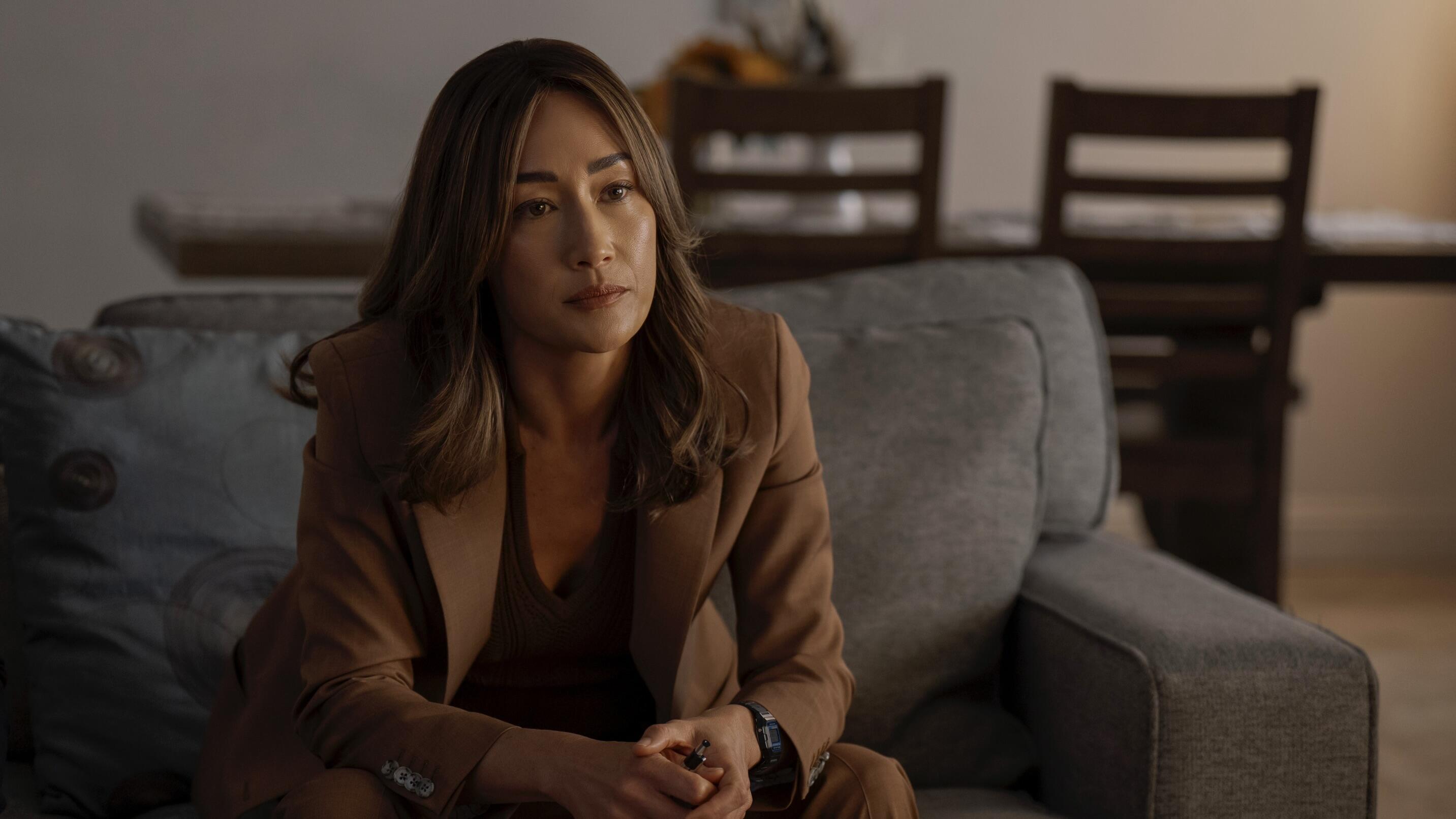 Maggie Q wears a brown pant suit and sits on a grey couch in a production still from Ballard.