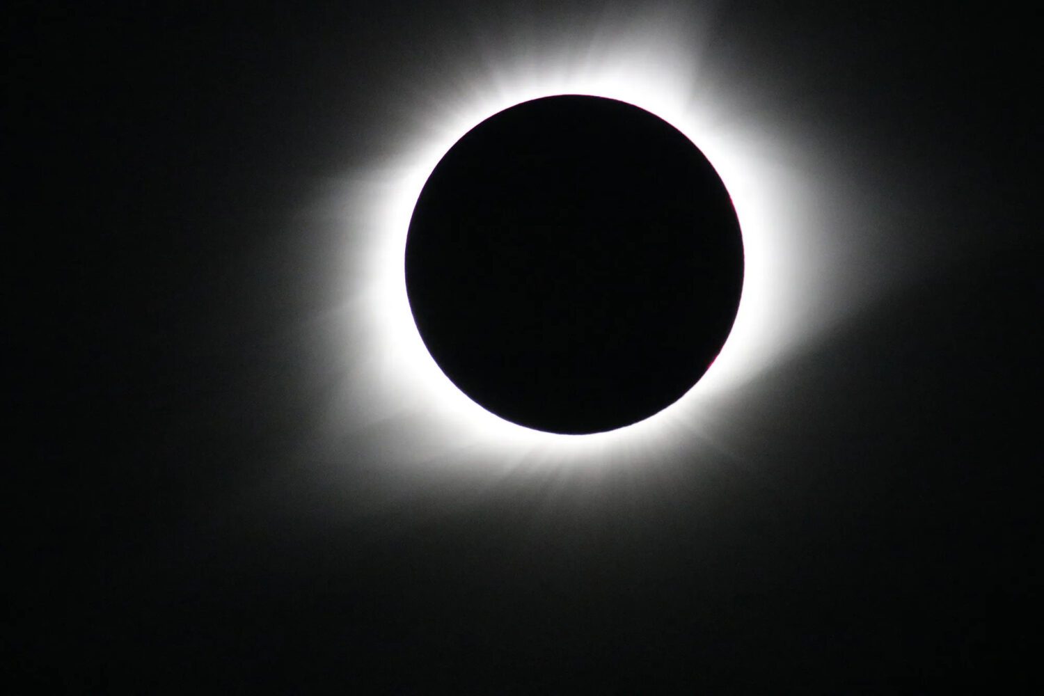 A stark, high-contrast photograph captures a solid black circle perfectly centered against a jet-black background. A bright, glowing ring of white light radiates outward from behind the dark circle, creating a soft, hazy halo with faint streaks extending into the surrounding darkness.