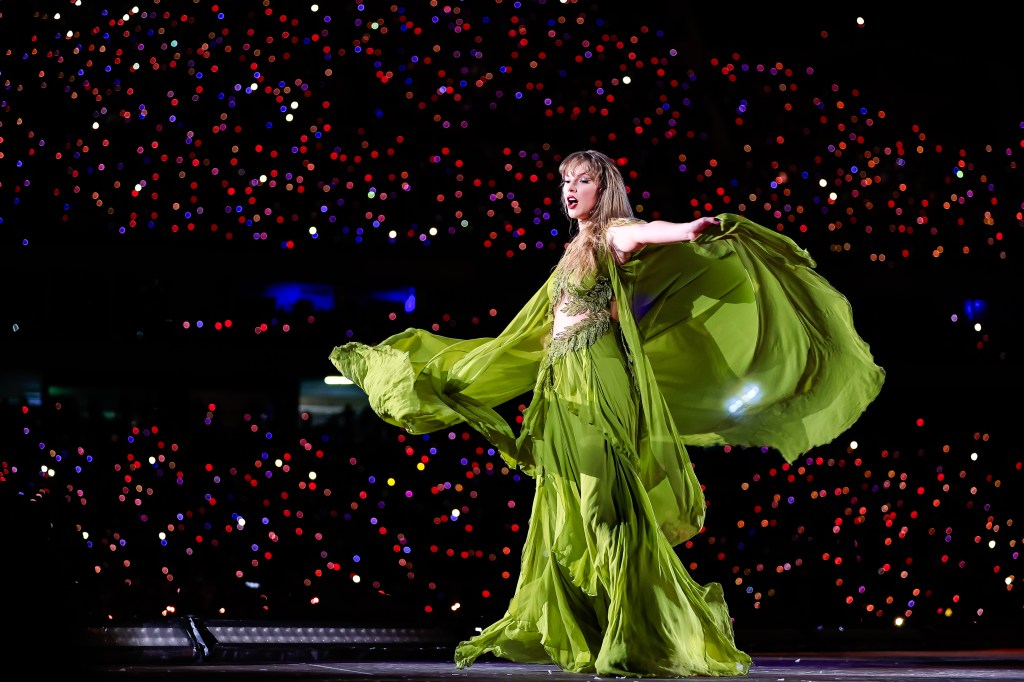 Taylor Swift performs onstage during "Taylor Swift | The Eras Tour" in a flowing green dress against a backdrop of glowing lights.