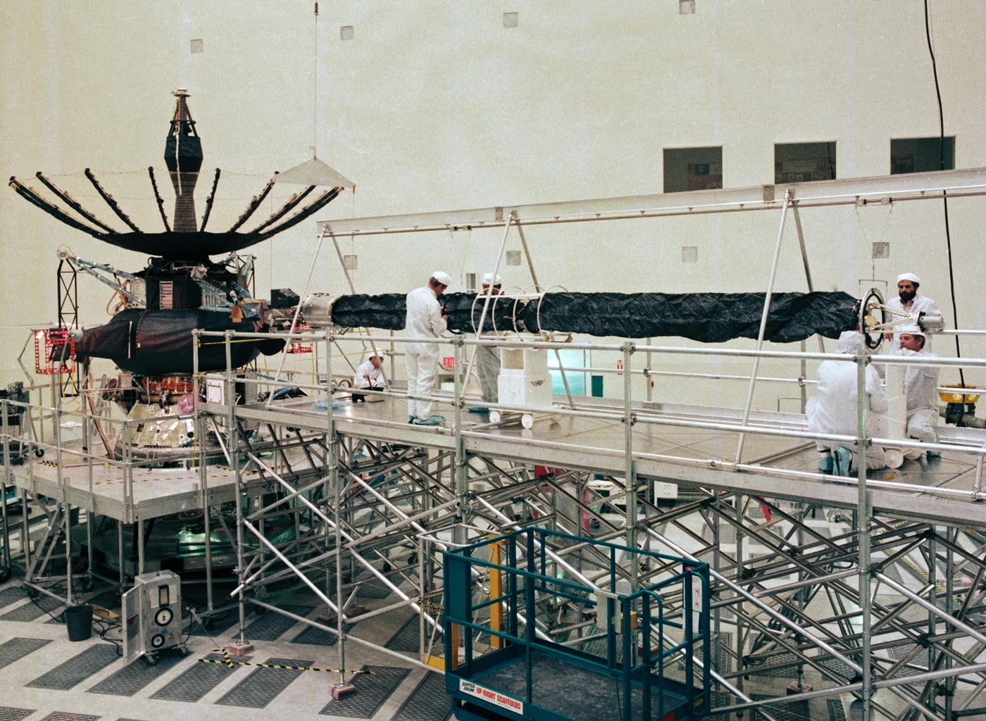 The Galileo spacecraft in a clean room at JPL in 1984, with its umbrella-like high-gain antenna fully opened above the spacecraft body and technicians in white gowns working on a long boom